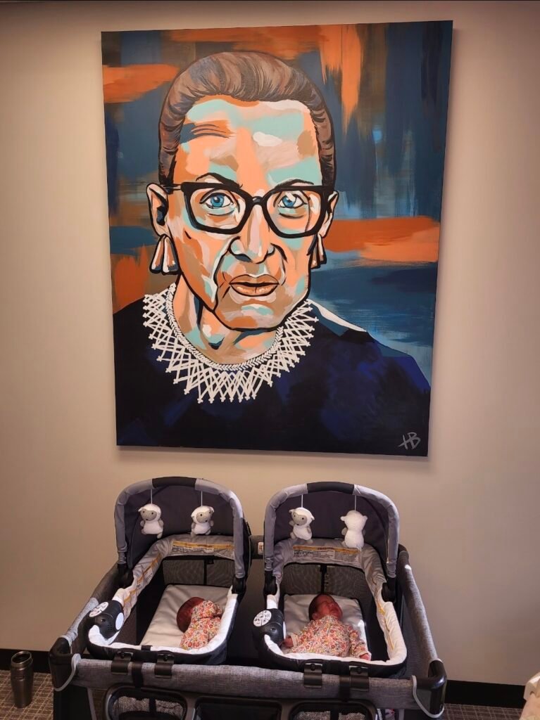 Sarah Lopez's two twin babies in a pack and play at work under a painting of Ruth Bader Ginsberg